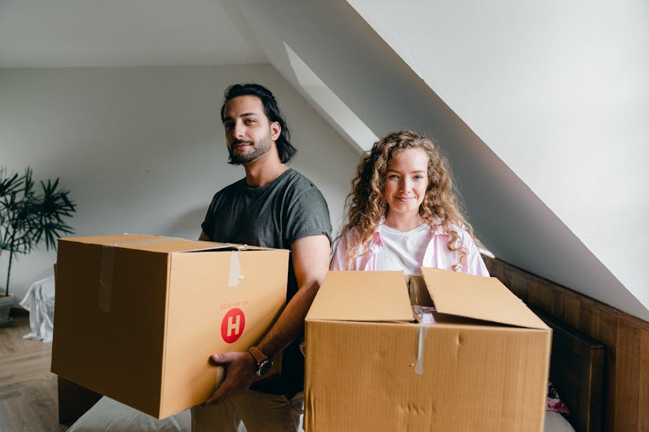 A man and a woman stand inside a residential room under a sloped ceiling, each holding a cardboard box as part of a home relocation process. The man, with dark hair and a beard, is wearing a grey t-shirt and is holding a large cardboard box with a red 'H' logo, indicating packing and furniture transport. The woman, with curly blonde hair, is dressed in a white top with a pink garment draped over her shoulders, and is carrying an open cardboard box filled with packing materials, possibly plastic wrap and paper. They are positioned near a wooden headboard, with a white wall and a potted plant in the background, suggesting indoor furniture handling. The lighting is natural, coming from an unseen light source, highlighting the details of the boxes, the packing materials, and the casual attire of the movers. The scene captures the loading process involved in professional removals, emphasizing the careful handling and packing typical of house removals services facilitated by Harringay Movers, reflecting effective packing and furniture transport during a home move.