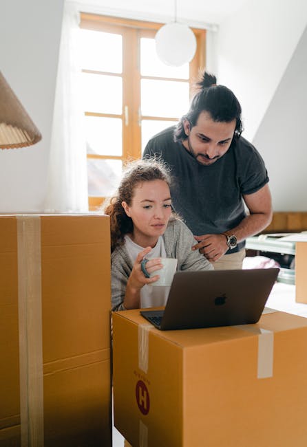 Inside a bright, well-lit room with large windows allowing natural light to fill the space, a man and a young girl are engaged in home relocation activities. The girl is seated at a table, holding a ceramic mug, while looking at a laptop with a focused expression. The man, standing behind her, leans over to observe the screen, with his hands resting on several cardboard boxes that are part of the packing process. The boxes, sealed with packing tape, are stacked in the room and positioned near the table to facilitate the loading process. The background features a wooden window frame with light curtains, a hanging ceiling light, and part of a desk or sideboard. This scene captures a moment of furniture transport and packing during a house move, with visible packing materials, cardboard boxes, and equipment indicative of a professional removal service such as Harringay Movers. The environment reflects a domestic setting preparing for a home relocation.