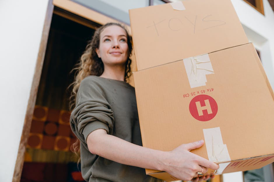 A man and a woman stand inside a residential room under a sloped ceiling, each holding a cardboard box as part of a home relocation process. The man, with dark hair and a beard, is wearing a grey t-shirt and is holding a large cardboard box with a red 'H' logo, indicating packing and furniture transport. The woman, with curly blonde hair, is dressed in a white top with a pink garment draped over her shoulders, and is carrying an open cardboard box filled with packing materials, possibly plastic wrap and paper. They are positioned near a wooden headboard, with a white wall and a potted plant in the background, suggesting indoor furniture handling. The lighting is natural, coming from an unseen light source, highlighting the details of the boxes, the packing materials, and the casual attire of the movers. The scene captures the loading process involved in professional removals, emphasizing the careful handling and packing typical of house removals services facilitated by Harringay Movers, reflecting effective packing and furniture transport during a home move.