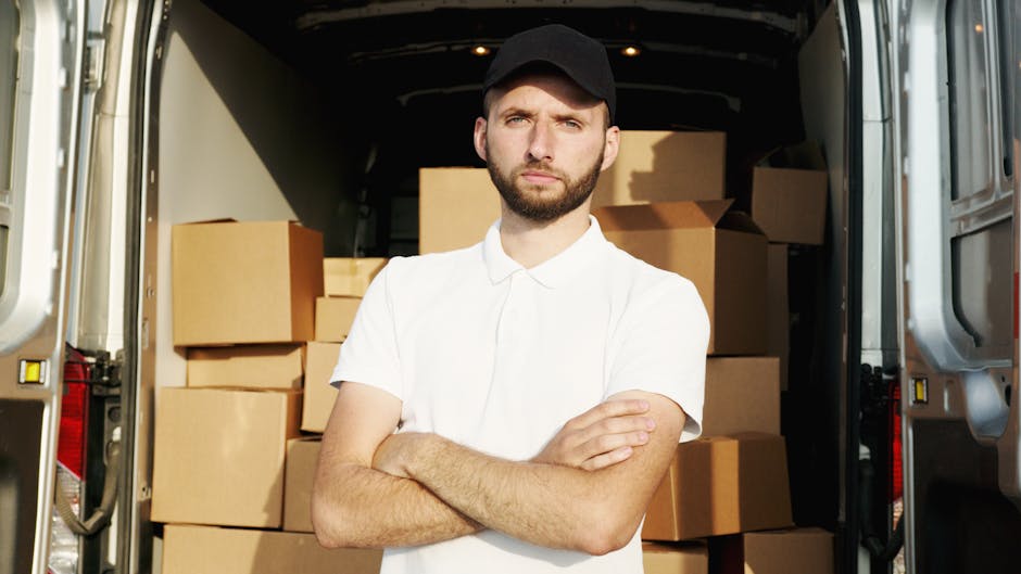 A young man with a beard and serious expression, dressed in a white polo shirt and black cap, stands inside the open rear doors of a moving van filled with neatly stacked cardboard boxes of various si