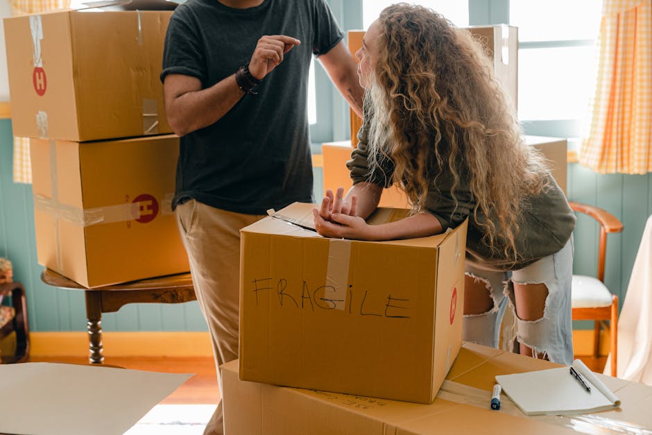 A young man with a beard and serious expression, dressed in a white polo shirt and black cap, stands inside the open rear doors of a moving van filled with neatly stacked cardboard boxes of various si