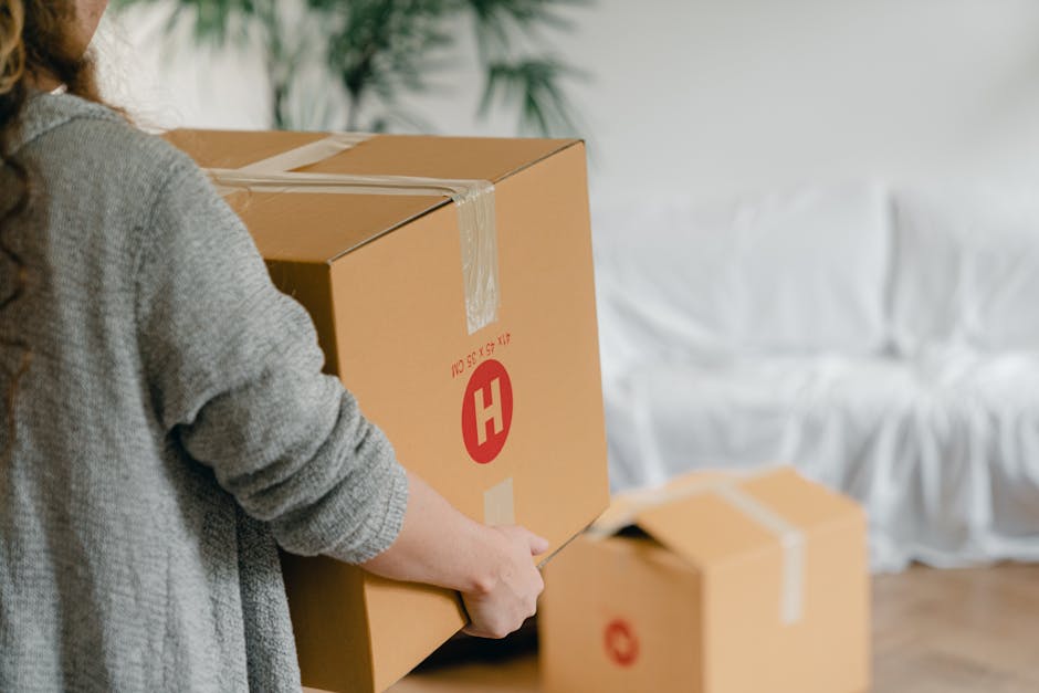 A person in a grey sweater is seen preparing to move a large cardboard box, which is sealed with packing tape and features a red 'H' logo, indicating it is part of a home relocation. The individual is holding a utility knife, likely to open or seal the box further. In the background, another smaller cardboard box with similar labeling sits on a wooden floor, near a white sofa covered with a protective fabric or slipcover. The scene appears to be indoors, inside a residential property, with soft lighting illuminating the space. The setting suggests a packing or loading process that is part of furniture transport or packing and moving activities, managed by Harringay Movers, a company specialising in removals and urgent same-day bookings in Harringay.