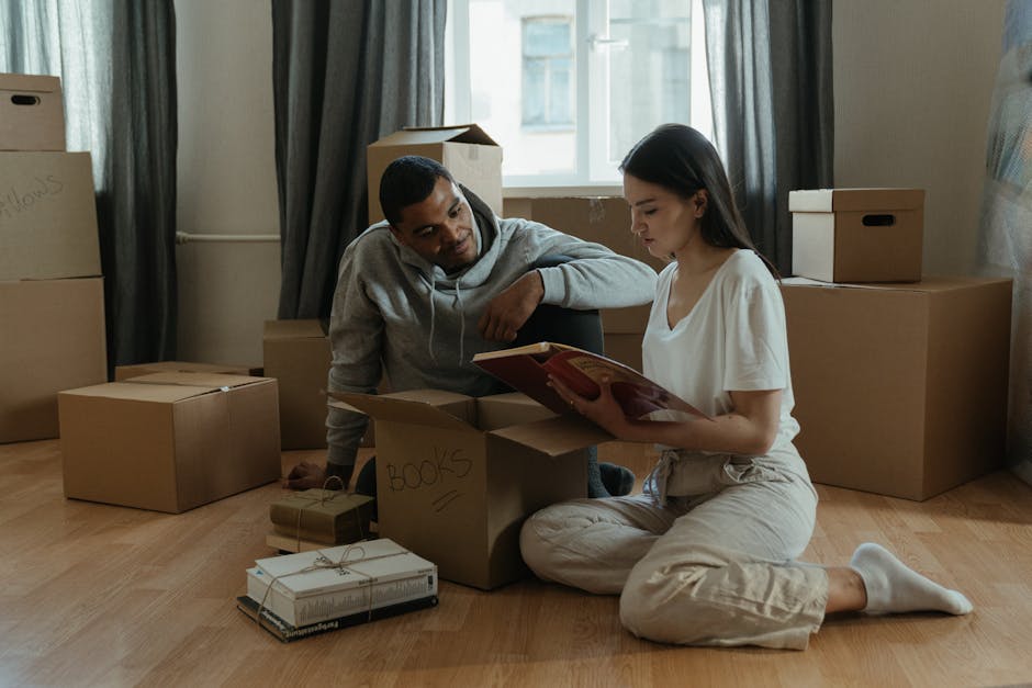 A person lying on the floor amidst a pile of cardboard boxes of various sizes, some sealed with packing tape and others open or partially collapsed. The boxes are stacked and scattered near a window with natural daylight illuminating the room. Visible are plastic handles on some boxes, indicating they are used for home relocation and furniture transport. The setting suggests an ongoing packing and loading process as part of a house move, with the person's legs outstretched behind the boxes. The scene reflects the organized chaos typical of packing for an emergency same day move, with furniture and contents prepared for transport by a professional removals service such as Harringay Movers. Equipment like packing materials, but no tools or vehicles, are visible, emphasizing the interior packing stage of the moving process.