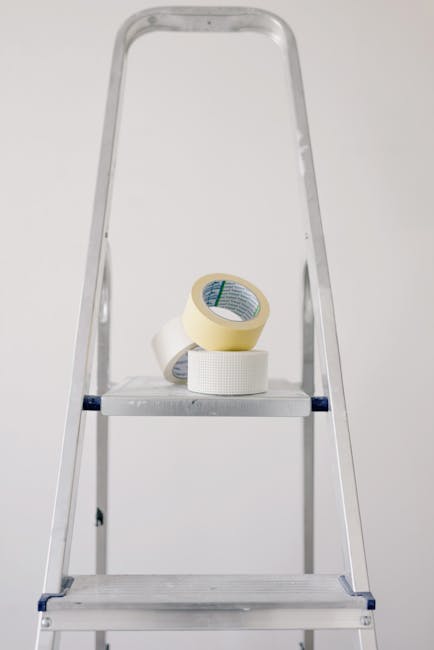 A close-up view of a silver aluminium step ladder with three steps, positioned indoors against a plain white wall. On the second rung from the top, there are three rolls of adhesive tape: one large beige roll and two smaller white rolls, placed for easy access during packing or repair tasks. The ladder appears steady and ready for use, often part of a home relocation or packing process by professional removals services such as Harringay Movers. The environment is well-lit with soft, natural or artificial light, highlighting the metallic surface of the ladder and the textures of the tape rolls. The scene emphasizes tools and materials commonly involved in furniture transport and packing during a move, supporting the context of house removals with narrow access challenges in areas like the Harringay Ladder, HARRINGAY. Appropriate for accessibility and SEO, the description focuses on the ladder's features and packing materials relevant to professional moving logistics.