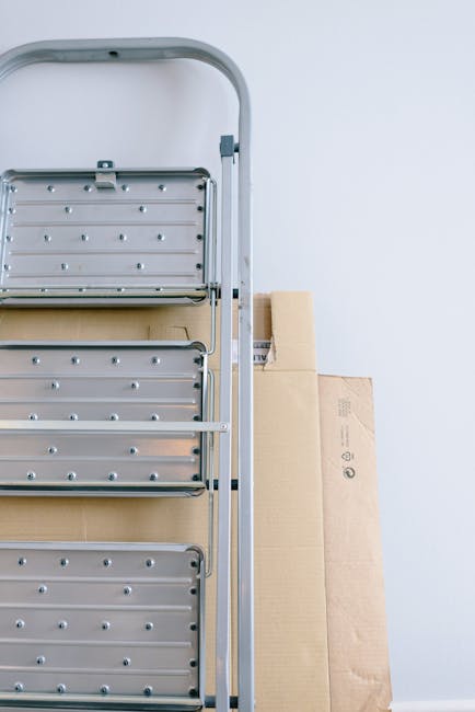 A close-up view of a silver aluminium step ladder with three steps, positioned indoors against a plain white wall. On the second rung from the top, there are three rolls of adhesive tape: one large beige roll and two smaller white rolls, placed for easy access during packing or repair tasks. The ladder appears steady and ready for use, often part of a home relocation or packing process by professional removals services such as Harringay Movers. The environment is well-lit with soft, natural or artificial light, highlighting the metallic surface of the ladder and the textures of the tape rolls. The scene emphasizes tools and materials commonly involved in furniture transport and packing during a move, supporting the context of house removals with narrow access challenges in areas like the Harringay Ladder, HARRINGAY. Appropriate for accessibility and SEO, the description focuses on the ladder's features and packing materials relevant to professional moving logistics.