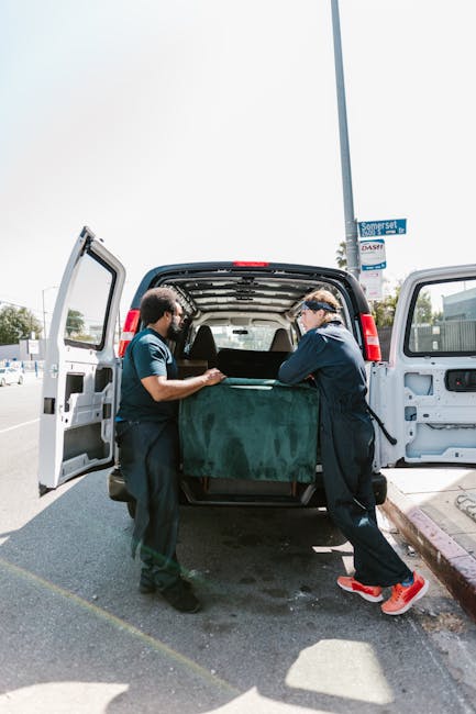 Two professional movers from Harringay Movers are engaged in the loading process of a home relocation, standing at the rear of an open cargo van on a paved street. One mover, dressed in navy work unif