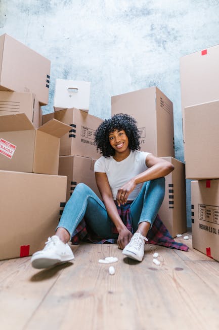 A young woman with curly black hair, wearing a white t-shirt, blue jeans, and white sneakers, is sitting on a wooden floor surrounded by multiple packed cardboard boxes in various sizes, some labeled with 'fragile' stickers and barcodes. She is smiling and appears relaxed after a home relocation process. The boxes are sealed with packing tape and stacked against a plain, light blue wall with a slightly textured surface. Some packing materials, such as small white packing peanuts, are scattered on the floor near her. The scene suggests she is in the midst of moving home, with items prepared for transport. This image exemplifies furniture transport and packing aspects of residential removals, aligning with services offered by Harringay Movers as featured on the 'Removals Green Lanes Harringay N4' page of harringaymovers.co.uk.