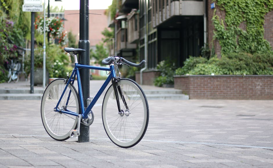 A blue bicycle with a black saddle and drop handlebars is locked to a black metal post on a paved sidewalk in an urban residential area. The bicycle appears to be in good condition, with thin tires and a minimalist frame design. In the background, there are steps leading to a building entrance, a low brick wall with greenery, and multi-storey residential buildings with balconies and external staircases, some covered in ivy. The scene is well-lit with natural daylight, and there are some blurred trees and additional bicycles further along the street. This setting exemplifies a typical urban environment where home relocation and packing may take place, with Harringay Movers providing local removal services in the area.