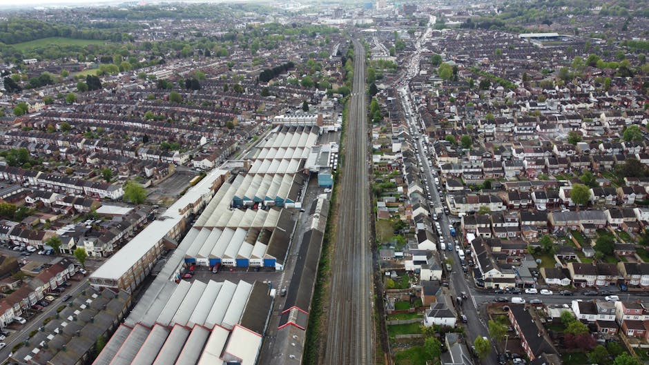 An aerial view of a densely populated residential area with rows of terraced houses on both sides of a railway line. The railway tracks run centrally through the image, extending from the foreground to the background, with a train station featuring multiple platforms and a large industrial warehouse complex with a series of pitched, translucent roofs visible on the left side. adjacent to the warehouse are loading docks with various trucks and containers. The surrounding houses are mainly brick-built with small gardens, and streets running perpendicular and parallel to the railway are filled with parked cars and some moving vehicles. In the distance, patches of green fields and trees can be seen, indicating the outskirts of the town. This image contextually relates to house removals and moving logistics, emphasizing transport and packing considerations for home relocation, as seen through the detailed depiction of building and transport infrastructure. Harringay Movers’ services are referenced through the overall relevance of moving and packing processes established by this scene.