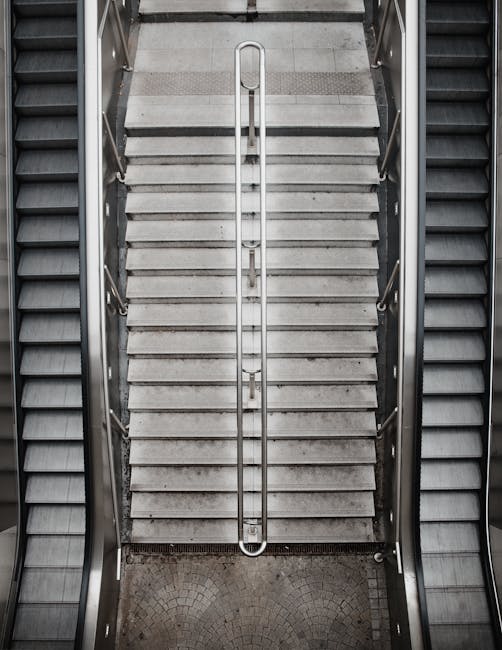 An overhead view of a concrete staircase with metal handrails on both sides, ascending between two sets of escalators in a building interior. The stairs appear slightly worn, with visible dirt and discoloration. At the bottom of the staircase, there is a semi-circular tiled landing area. The surrounding area is illuminated by ambient lighting, highlighting the utilitarian design of the stairs and escalators. This environment suggests a public space or transport hub where moving and logistics services such as assistance with stairs access problems are often required, which Harringay Movers can potentially support as part of their relocation solutions including furniture transport and home removals involving stairs access challenges.