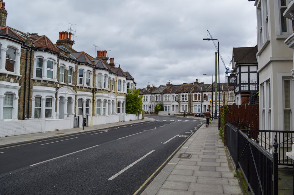 A residential street scene with white terraced houses featuring bay windows, brick facades, and red-tiled roofs, under an overcast sky. The pavement runs along the right side of the image, with a black metal fence and some plants visible in front of the houses. The road in the centre has marked lanes and a few parked cars at the far end. Streetlights and a crane are also visible in the background. The scene depicts a quiet area suitable for home relocation services, with no visible moving activity or furniture in the image but set in a typical UK neighbourhood environment, aligning with house removals and furniture transport contexts, as might be handled by Harringay Movers.