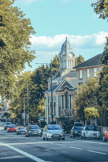 A busy street scene in Harringay with several parked and moving cars along the road. To the right, there is a building with a sloped roof and multiple windows partially obscured by green trees. Prominently visible in the background is a historic clock tower with a pointed spire, ornate architectural details, and a clock face showing the time, situated atop a church or civic building. The street features lampposts and traffic signage, and the sky above is clear with some scattered clouds. The scene captures an active urban environment during daylight hours, internally relevant for house removals or home relocation services such as those offered by Harringay Movers, with a focus on transportation logistics and cityscape context.