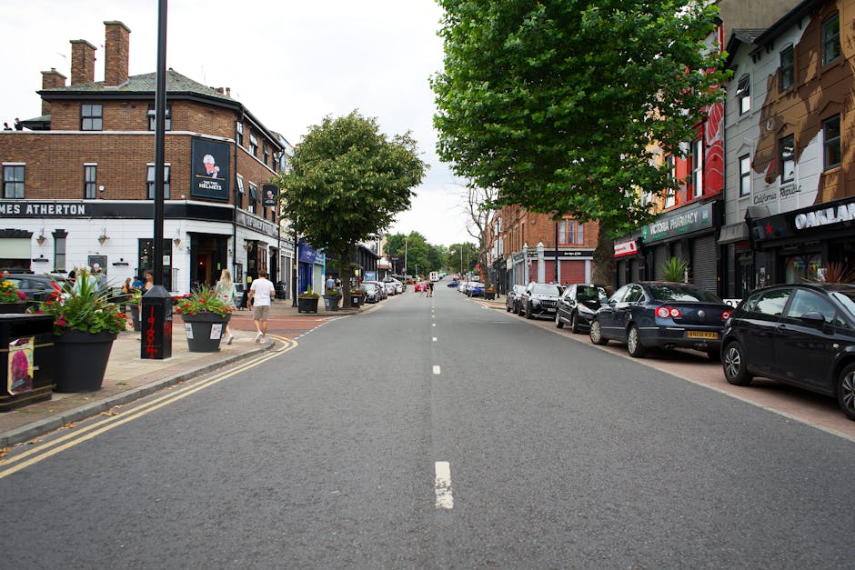 A residential street scene with white terraced houses featuring bay windows, brick facades, and red-tiled roofs, under an overcast sky. The pavement runs along the right side of the image, with a black metal fence and some plants visible in front of the houses. The road in the centre has marked lanes and a few parked cars at the far end. Streetlights and a crane are also visible in the background. The scene depicts a quiet area suitable for home relocation services, with no visible moving activity or furniture in the image but set in a typical UK neighbourhood environment, aligning with house removals and furniture transport contexts, as might be handled by Harringay Movers.
